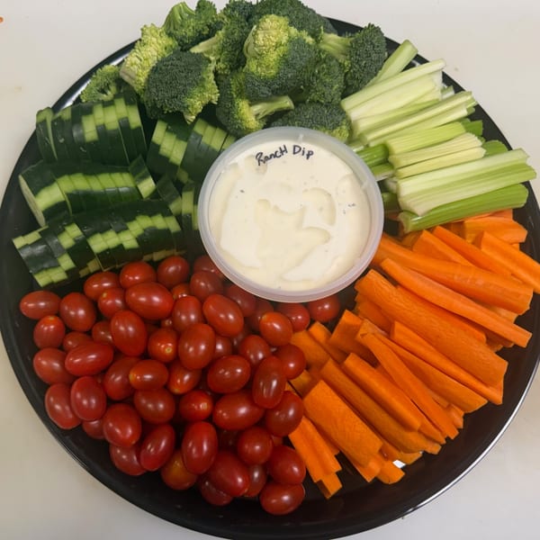 A black platter with broccoli, celery, cucumbers, cherry tomatoes, carrots, and a ranch dip.