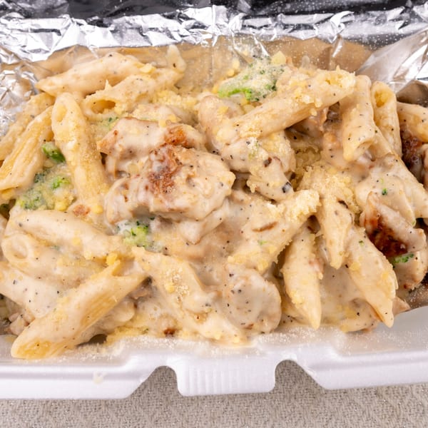 Creamy penne pasta with chicken and broccoli, topped with cheese, in a foil-lined takeout container.